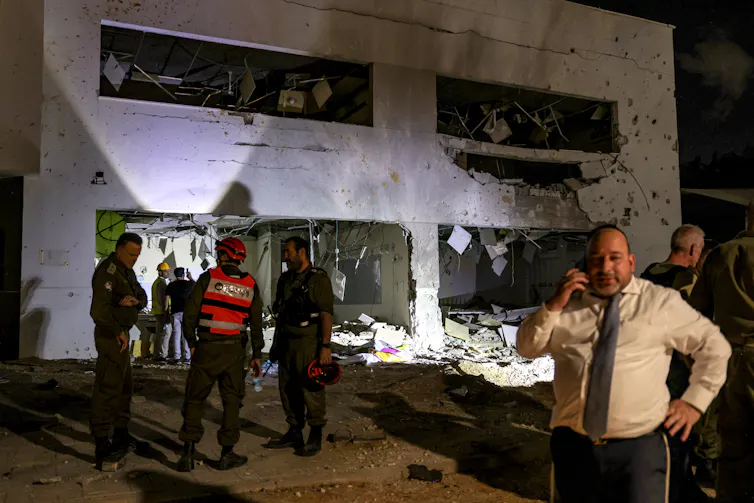 People stand around a gray building at nighttime that appears damaged.