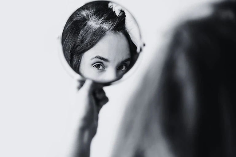 A woman inspects grey hairs in a circular mirror.
