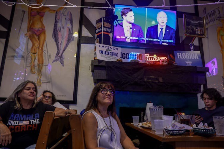 People at a restaurant watching a video screen up high on a wall.