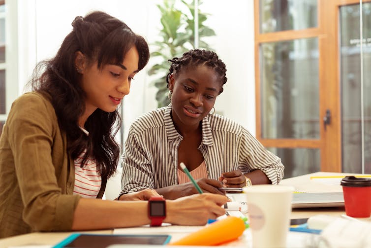 Two young women sit side by side at a desk with books and drinks.