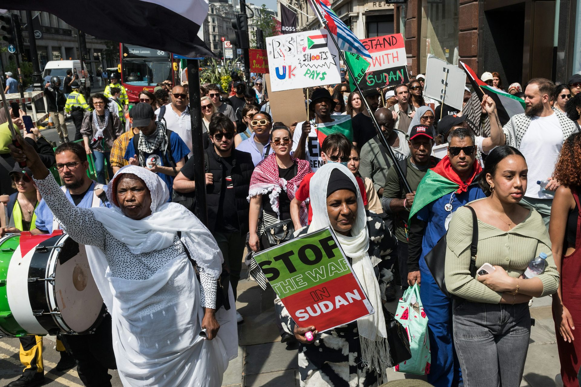 Women and men holding up placards written 'stop the war in Sudan' while holding red, green and white flags and drums