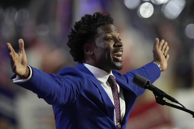 A Black man in a blue suit holds out his arms as he speaks into a microphone.