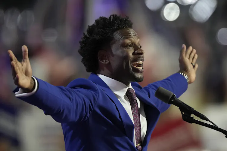 A Black man in a blue suit holds out his arms as he speaks into a microphone.
