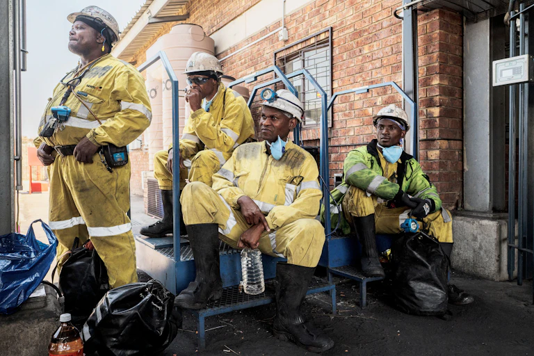 Four coal mine workers in yellow overalls and white hard hats wait to start a shift in a coal mine