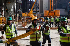 A dozen workers in yellow high visibility vests and hard hats are working on huge pieces of equipment at a power station