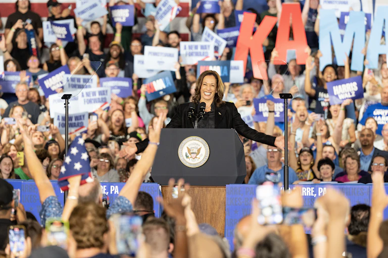 A woman stands on a stage at a lectern in front of a large crowd of people who hold blue and white signs and wave their hands.