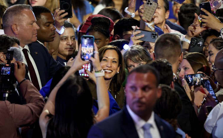 A woman stands in a crowd of people and smiles as others hold up phones and take photos of her.