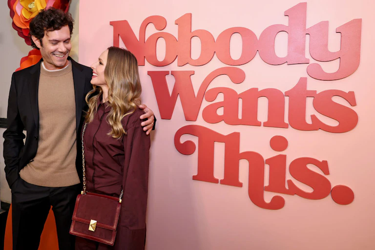 A man in a sweater and jacket and a woman in a dark red top pose in front of a sign that says 'Nobody Wants This' in red letters.