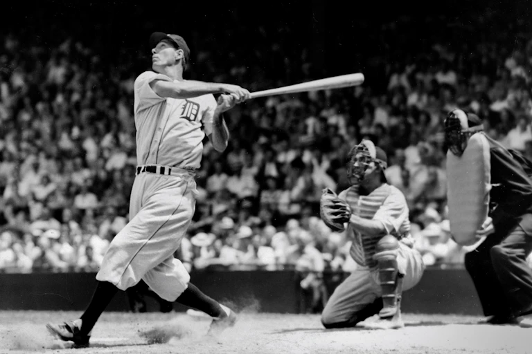 With the crowd in the background, a baseball player at the plate swings at the ball.
