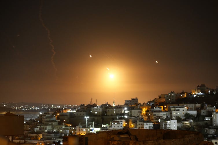 A glow is seen above some buildings with bursts of lights from missiles scattered around.