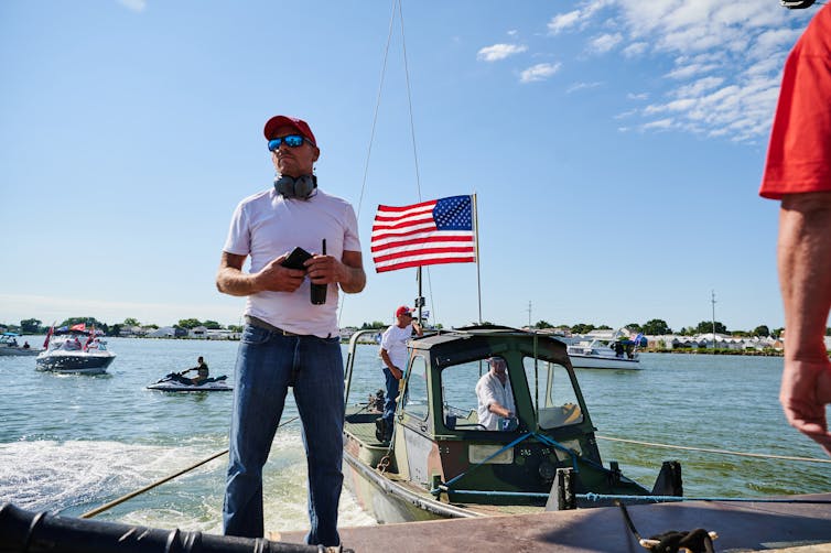 Man holding binoculars stands on a dock in front of a large body of water and a boat with American flag