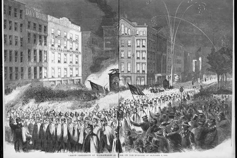A large procession of men in capes and hats, marching in a city while fireworks are set off.