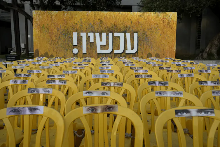 Rows of empty yellow chairs beneath the Hebrew word for 'now'.