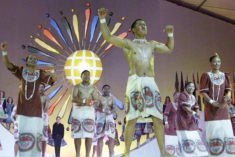 Men and women in traditional Samoan attire dance in from of the Commonwealth emblem.