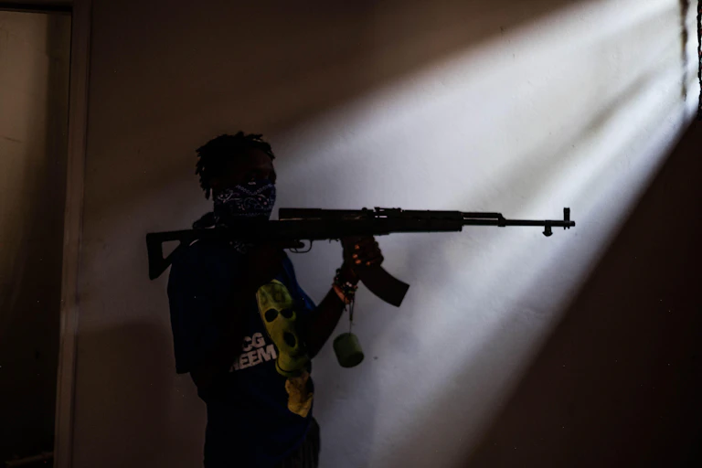 A man standing in a dark room in a pocket of light with an automatic rifle resting on his shoulder.