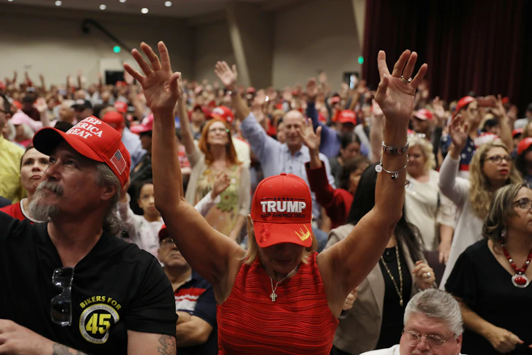 A large group of people, standing with their hands and eyes raised toward the ceiling. A few are wearing caps with 'Trump' and 'Make America Great Again' written on them.