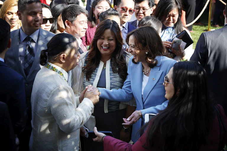 A woman wearing a light blue blazer shakes hands with a man with a ponytail and a beige jacket. She is surrounded by other people who are smiling.