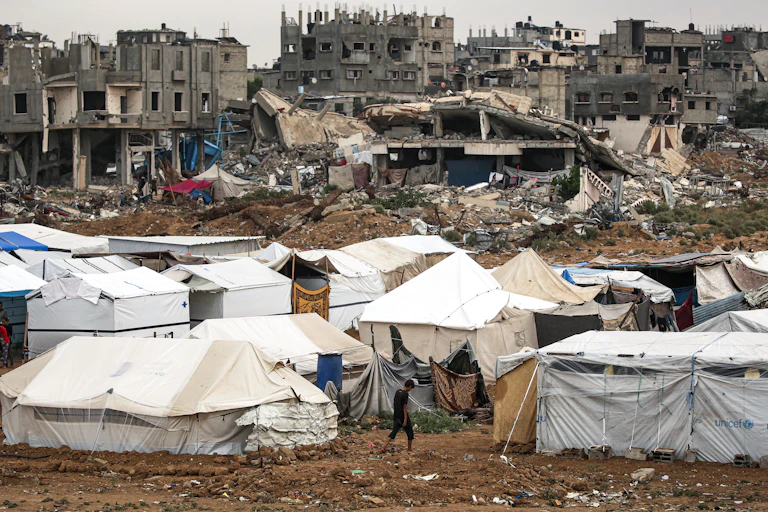 Tents and makeshift shelters are clustered in an open space between partially destroyed buildings.