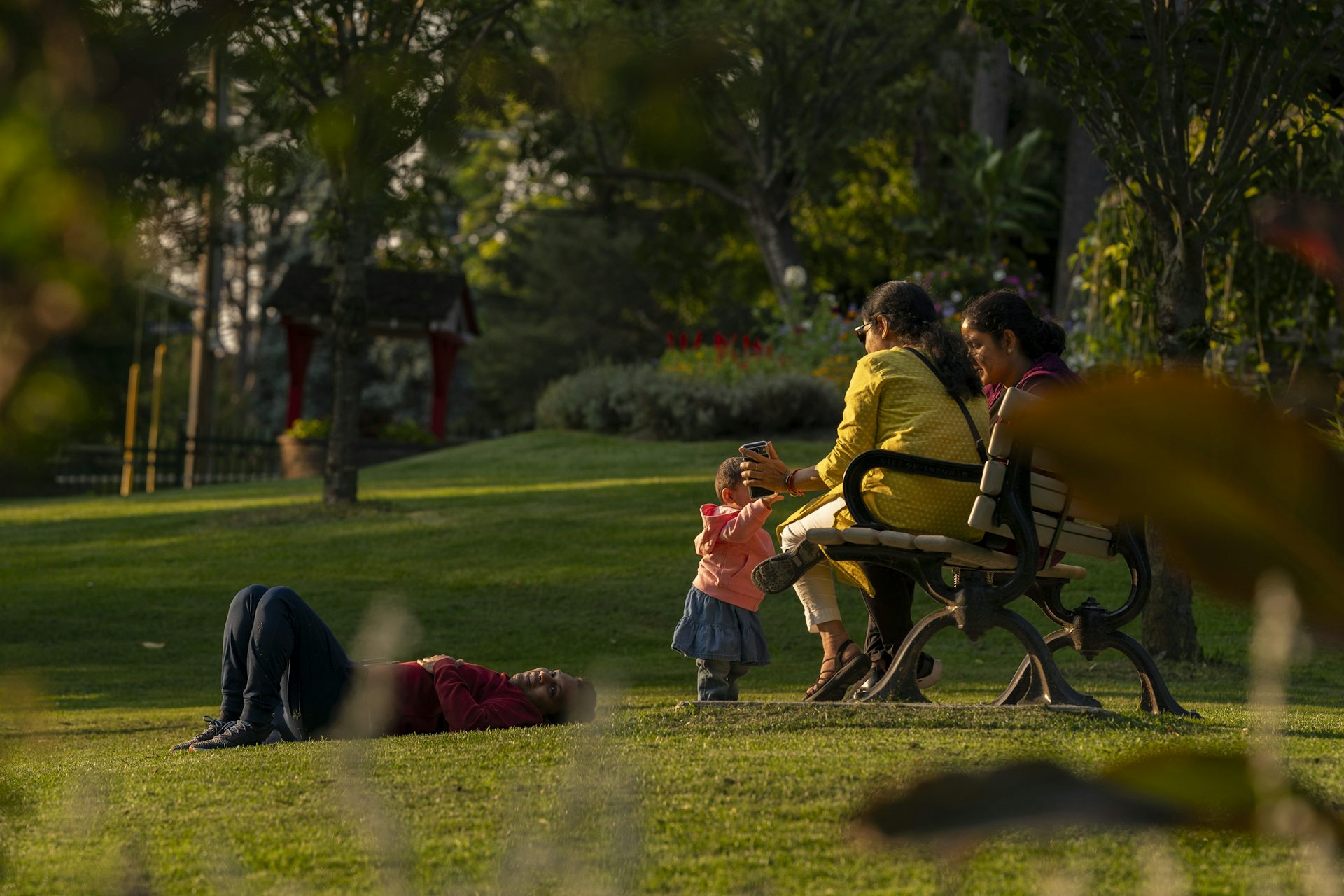 A child walks up to two women who are seated on a bench on a park. Beside them, a person lies on their back on the grass.