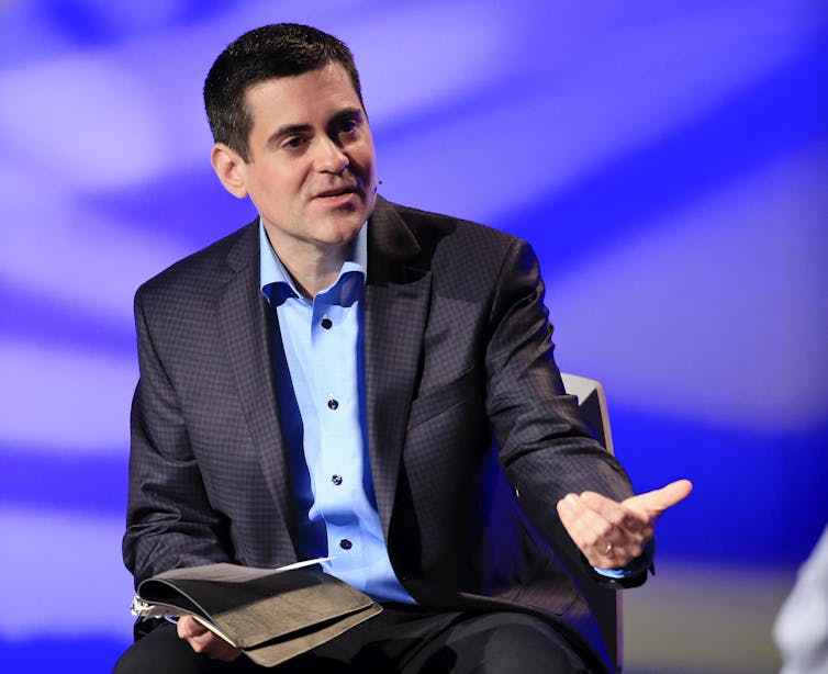 A man in a blue shirt and dark navy suit gestures with one hand while holding a book in another, as he speaks.