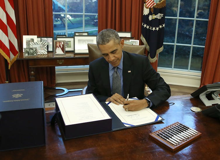 Man in suit and tie sits at large wooden desk and signs document