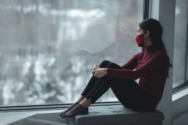 a woman wearing a surgical mask sits on a windowsill — outside there is snow on the ground