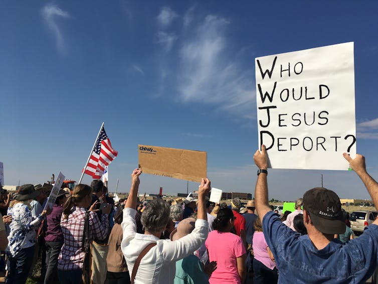 Several protestors stand while holding placards, one of which says, 'Who would Jesus deport.'