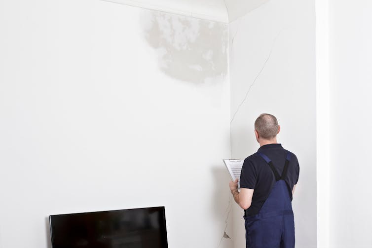 A man assessing a white wall with water damage and a large crack in the plaster.