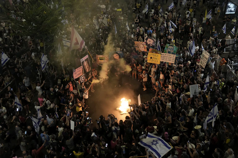 A crowd of people holding placards gather in a circle around a small fire at night.