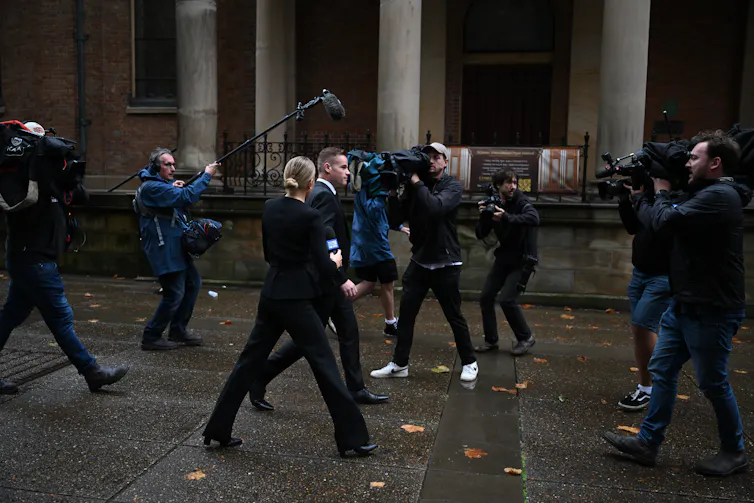 A man in a suit walks down a street surrounded by journalists and camera operators.