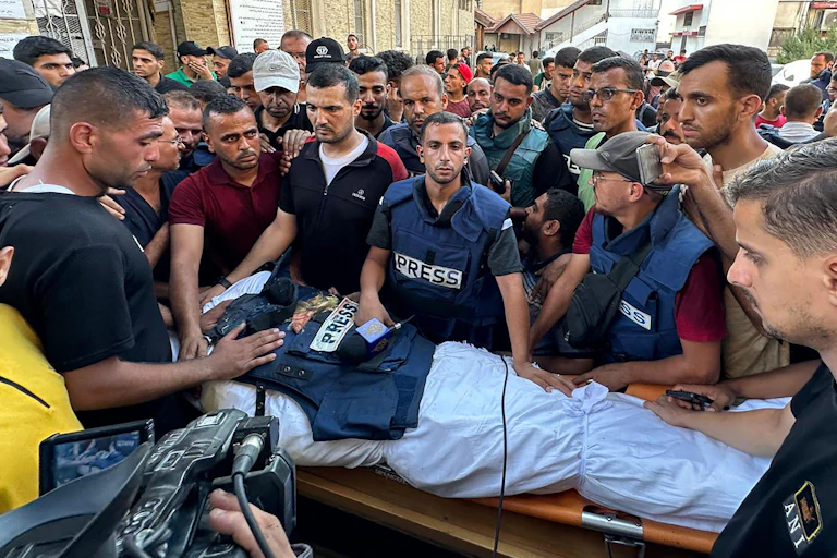 A crowd of men, some in press jackets, gathering around the sheet-wrapped body of al-Ghoul, which has a press flak jacket laid on top of it