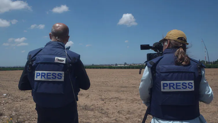 View from behind of two reporters in protective flak jackets reading 'press' pointing cameras towards the Gaza Strip