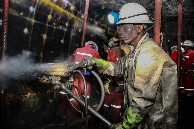 A man in a hard hat and a heavy uniform, his eyes protected by safety goggles, works underground in a mine