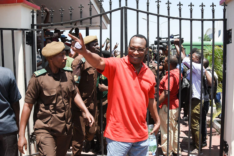 A man in a red shirt and wearing specs holds up two fingers in a wave while surrounded by soldiers in a brown uniform