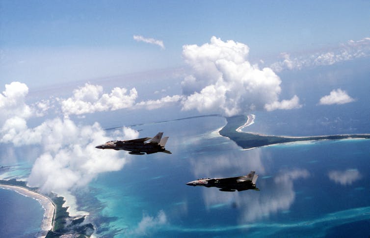 Two fighter jets flying over a tropical atoll.