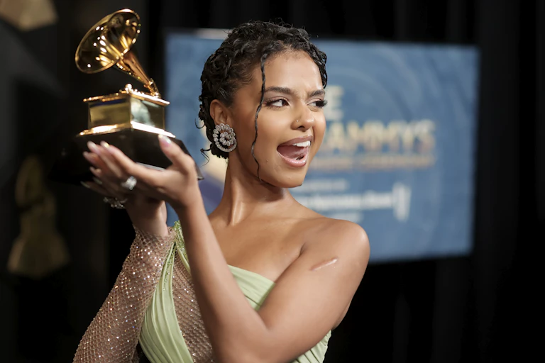 A young woman in an off-the-shoulder evening gown sticks her tongue out cheekily while displaying a large gold statue shaped like a gramophone
