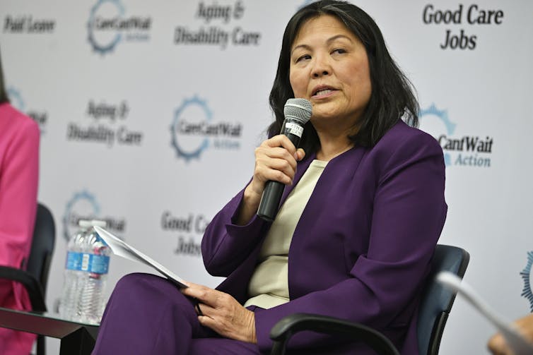 A woman in a purple pantsuit holds a microphone while speaking at a conference.
