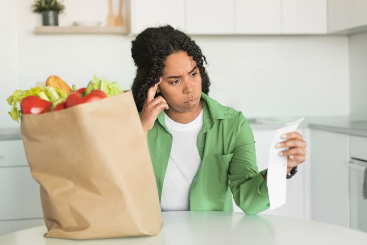 A woman sitting in a kitchen with a bag of groceries on the table, looking at the receipt