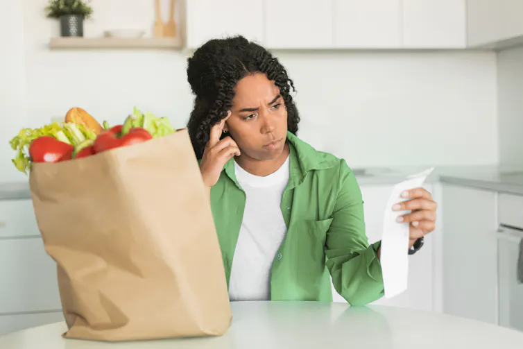 A woman sitting in a kitchen with a bag of groceries on the table, looking at the receipt