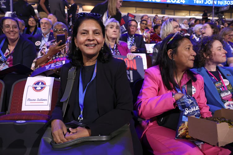 A woman smiles and sits next to other women in a crowded room with people, some of whom has signs that are blue and red.
