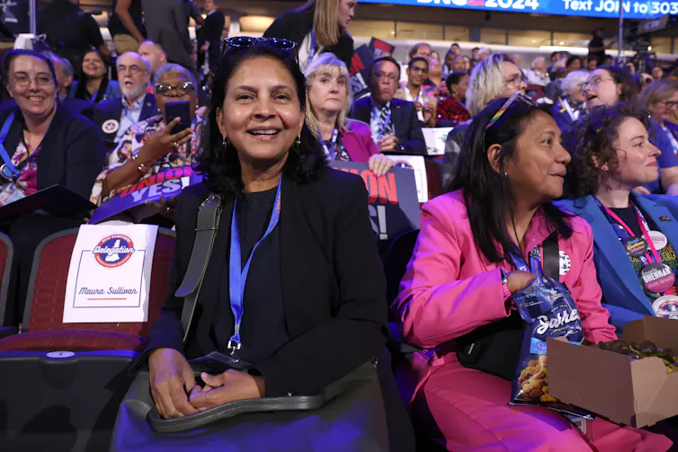 A woman smiles and sits next to other women in a crowded room with people, some of whom has signs that are blue and red.
