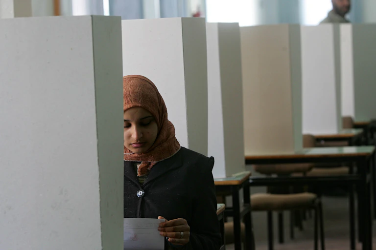 Uma mulher com um lenço na cabeça olha para uma folha de papel em um cubículo.