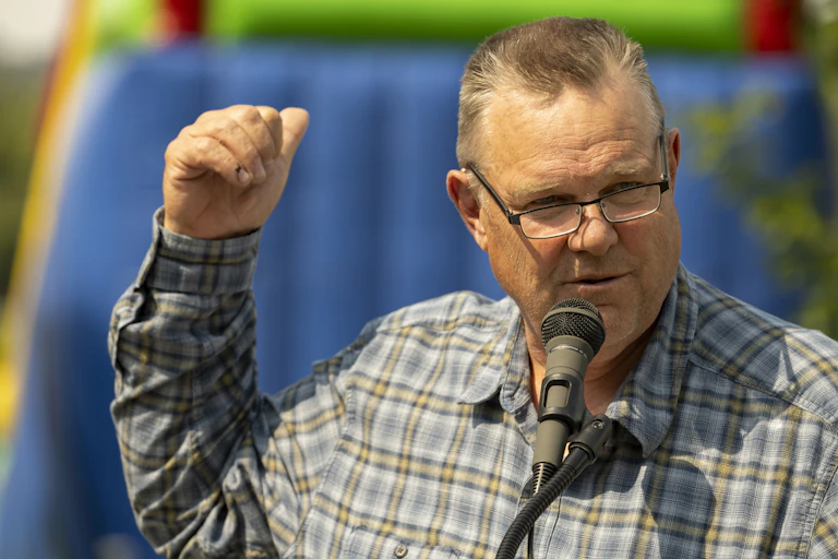 A man with a flattop haircut, wearing a plaid shirt, speaking into a microphone.