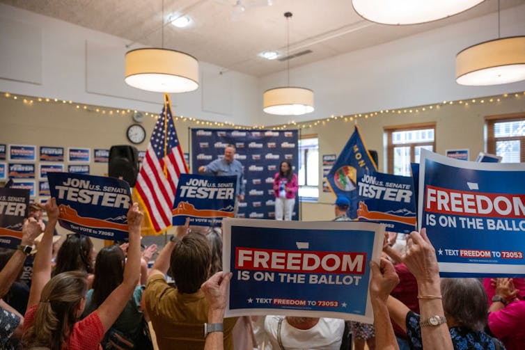 A man talking at the front of a room with people waving flags and signs that say 'FREEDOM is on the ballot.'