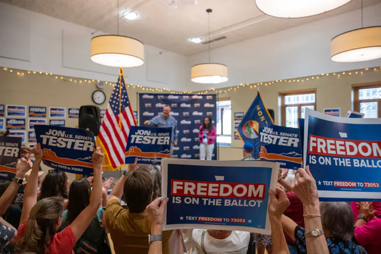 A man talking at the front of a room with people waving flags and signs that say 'FREEDOM is on the ballot.'