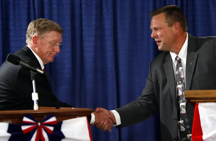 Two men shaking hands on a stage behind lecterns.