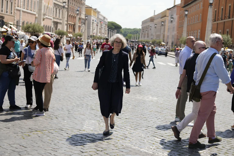 A woman with a black skirt suit, glasses and gray hair walks along a wide, cobbled street as other people stroll around her.