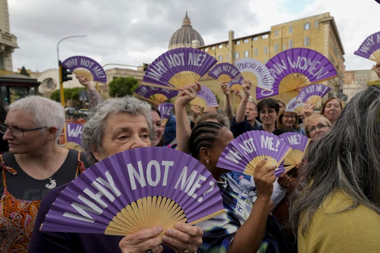 Women standing outdoors hold unfurled purple fans that say 'Why not me'?