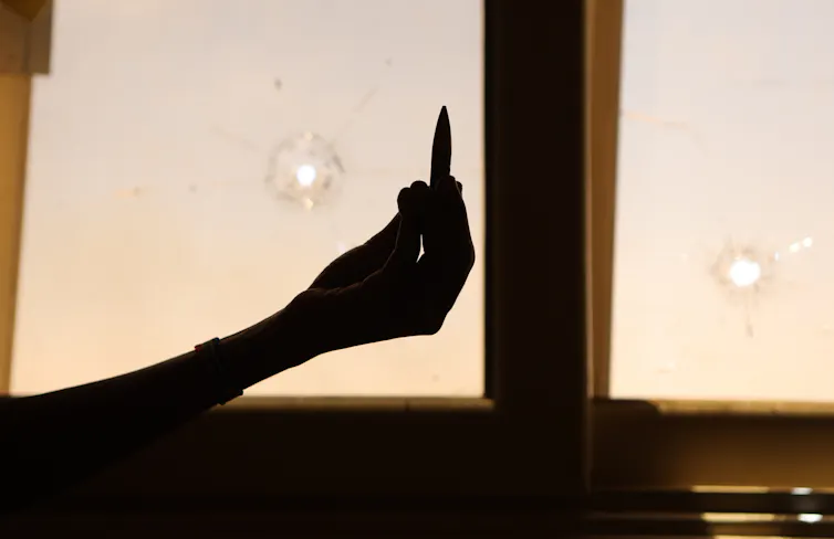 A person holding up an empty bullet case in front of a damaged window.
