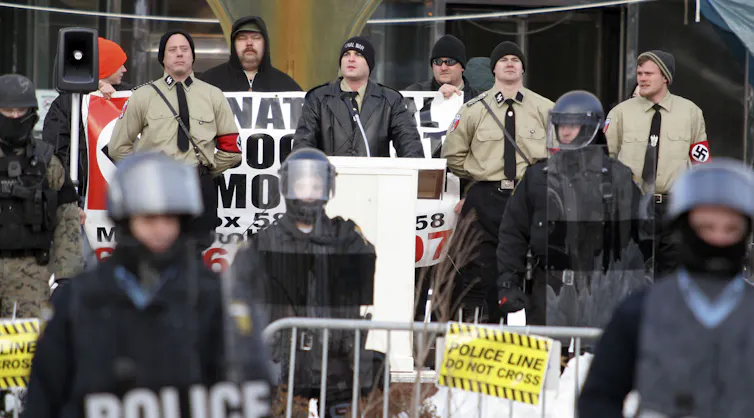 A group of men wearing brown shirts and Nazi swastikas stand in front of the Toledo, Ohio City Hall building and are surrounded by policemen.